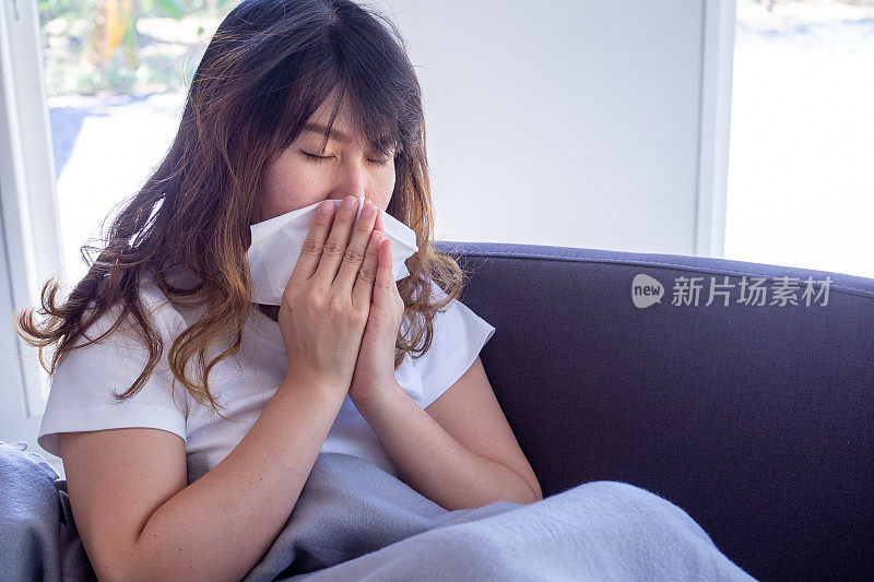 Long haired woman sitting on the sofa is suffering from flu, cough and sneezing. Sitting in a blanket because of high fever and cover their nose with tissue paper because sneezes all the time.